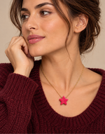 A close-up of a woman wearing a gold chain necklace with a bright pink star-shaped pendant, paired with a burgundy knit sweater against a warm beige background.