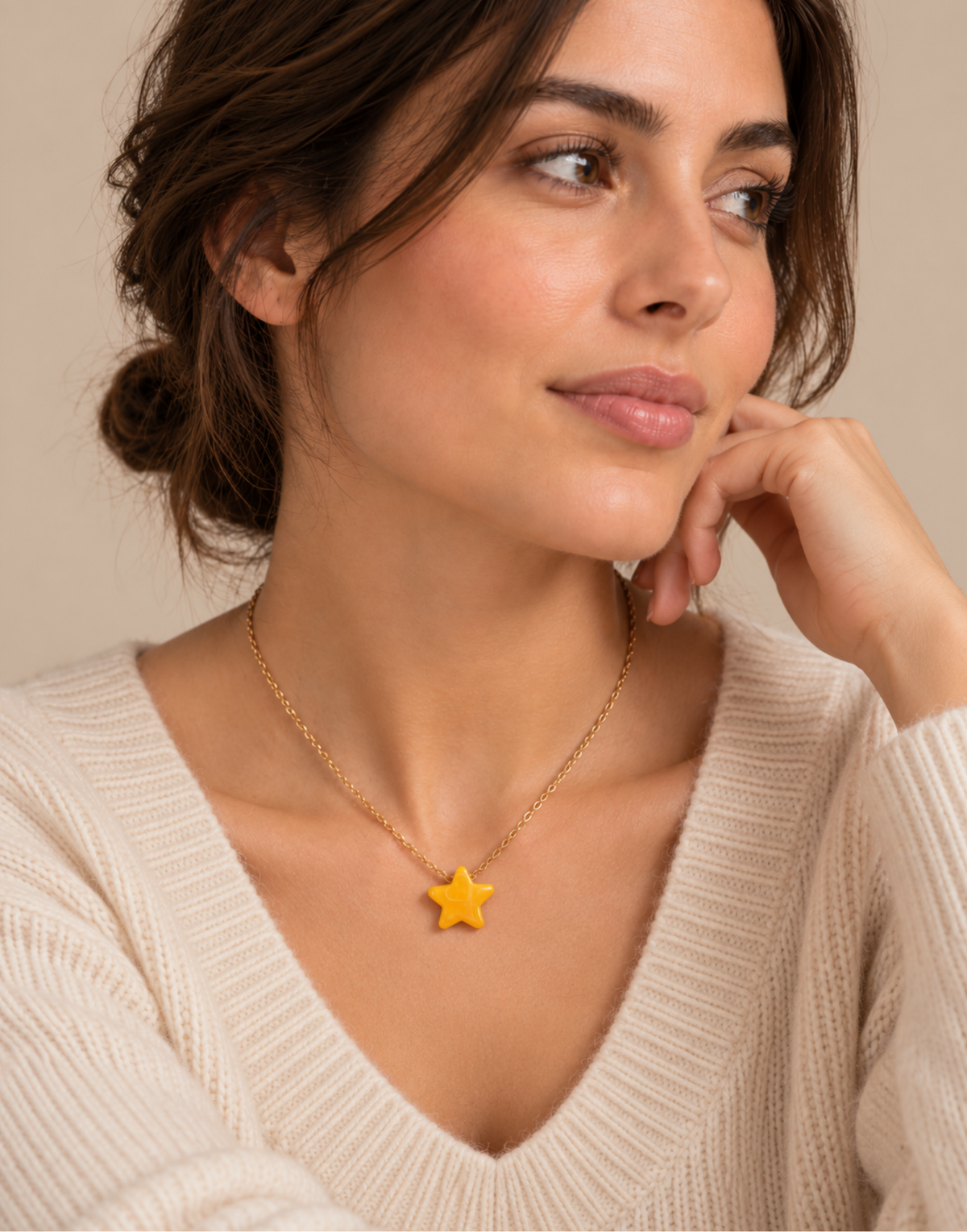 A woman wearing a delicate gold chain necklace with a small yellow star pendant, styled with a cream-colored knit sweater against a warm beige background.